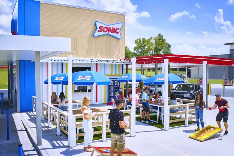 Customers enjoying food and beverages at the outdoor seating area of a Sonic drive-in franchise restaurant on a sunny day.